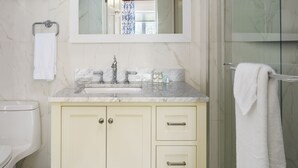 A bathroom with a marble-tiled wall, a glass shower enclosure, a vanity with a mirror, and a towel rack. - The Oxford House (Pembroke Parish)