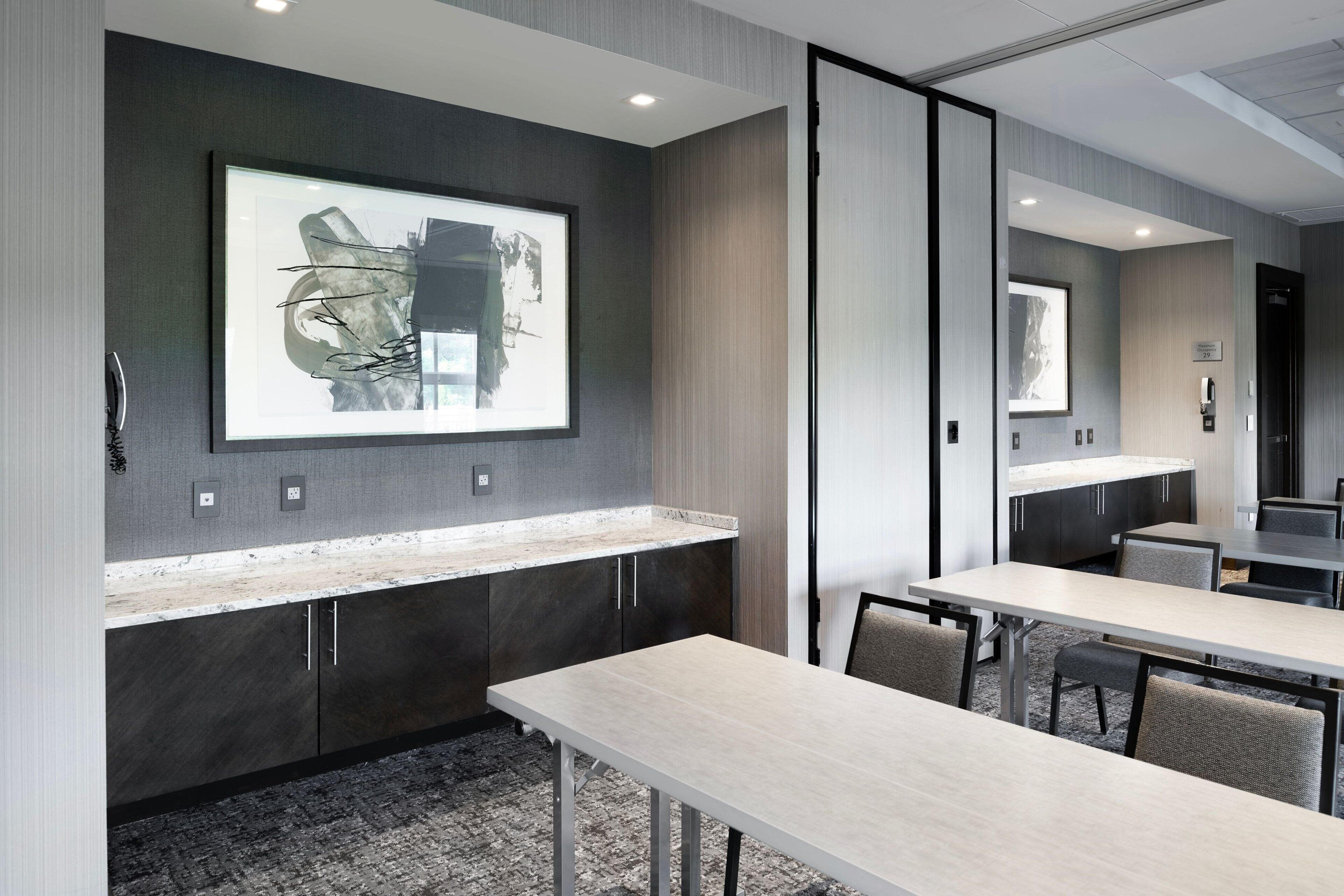 A modern conference room with a large table, chairs, a projector screen, and a kitchen area with cabinets and a countertop.