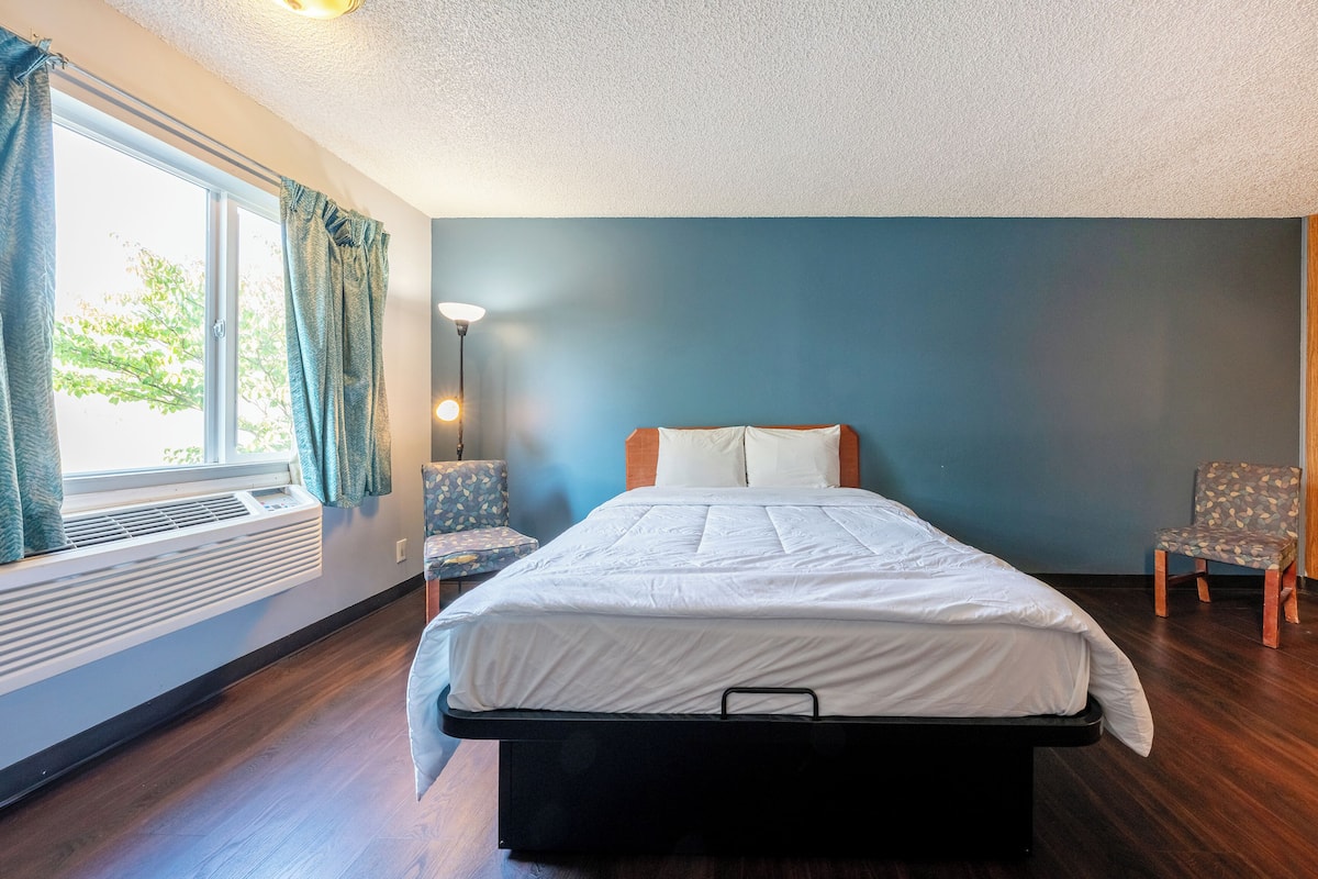 A bedroom with a bed, two chairs, a window with curtains, a wall-mounted heater, and a ceiling light.
