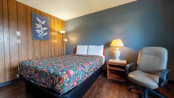 A hotel room with a wooden paneled wall, a bed with a colorful floral bedspread, a desk with a lamp, and a chair.