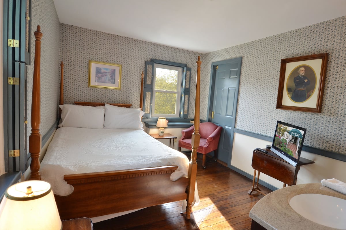 A bedroom with a four-poster bed, a desk with a chair, a window, and a bathroom with a sink and mirror.