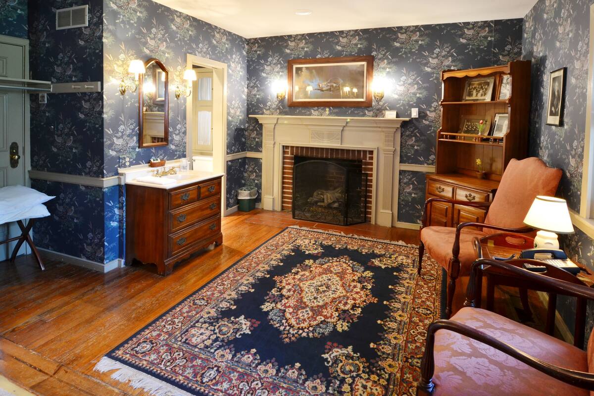 A room with a fireplace, wooden furniture, a patterned rug, and a mirror with wall sconces.