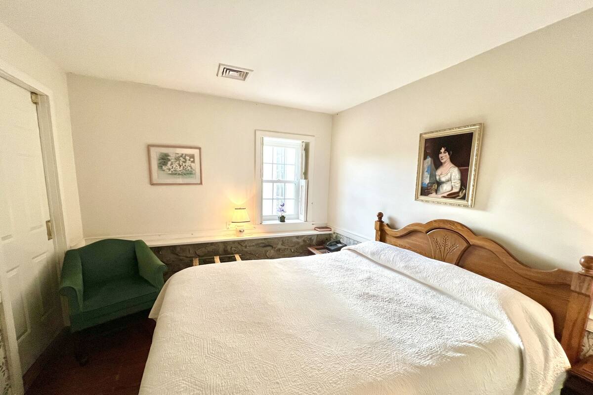 A bedroom with a large bed, a green chair, a window with a view, and a painting on the wall.