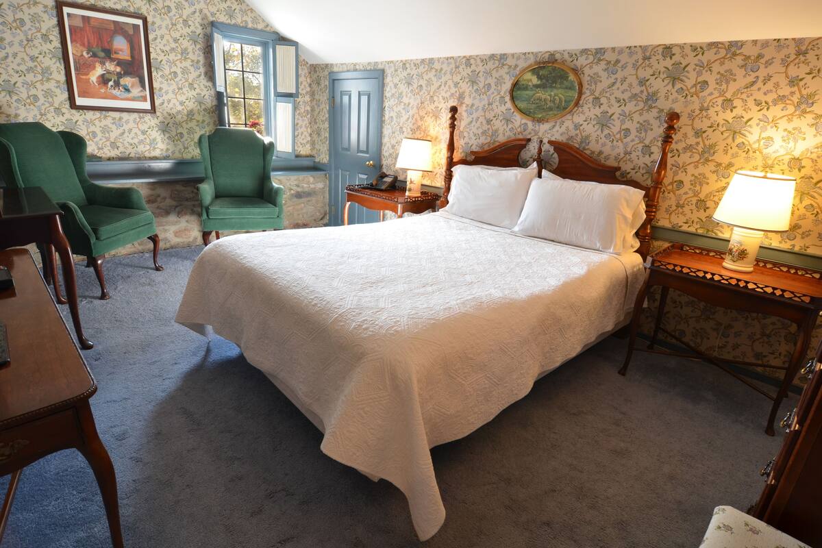A neatly arranged bedroom with a large bed, two chairs, a table, and a lamp.