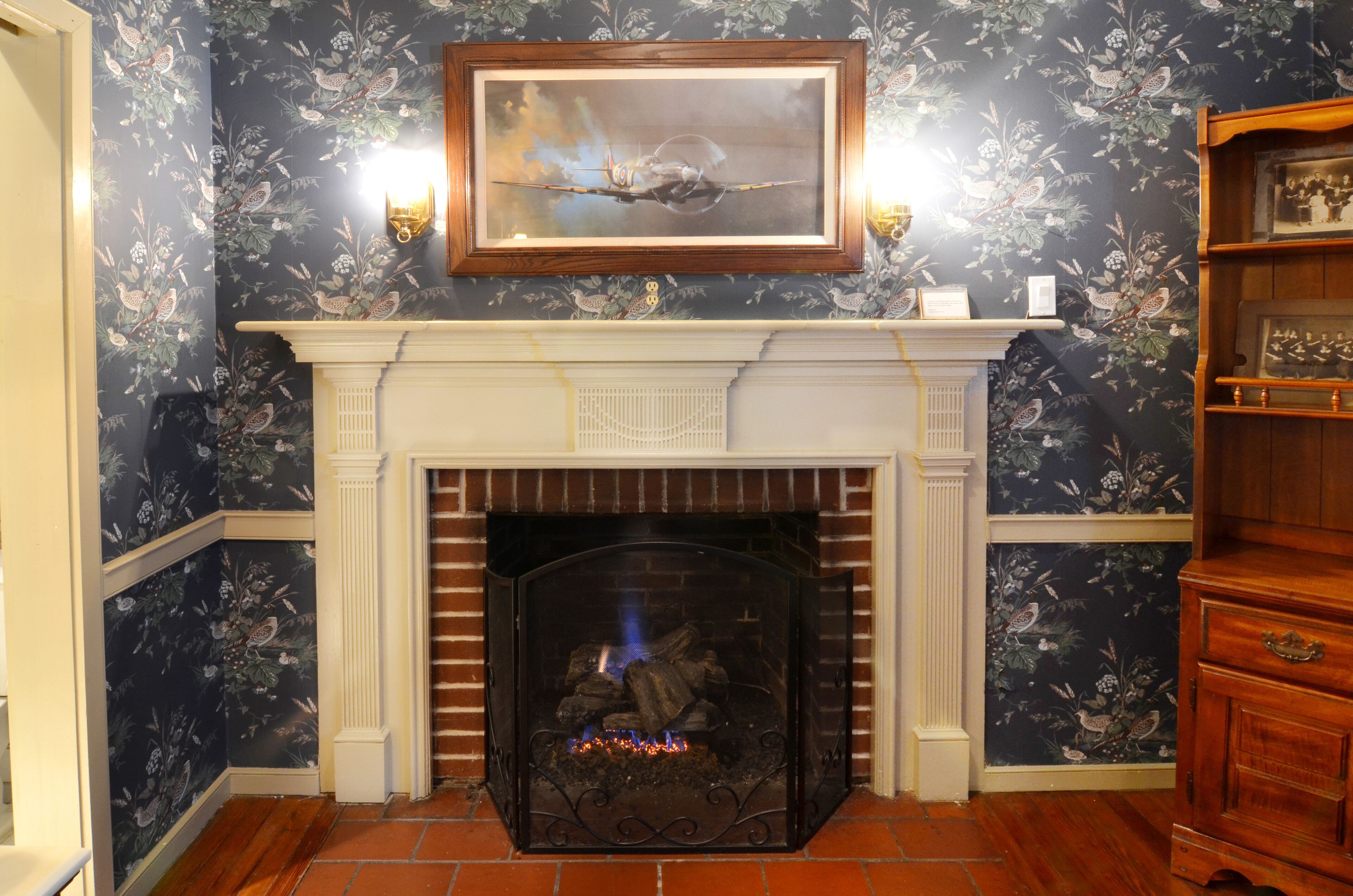 A fireplace with a mantel, a framed artwork above it, and a patterned wallpaper.