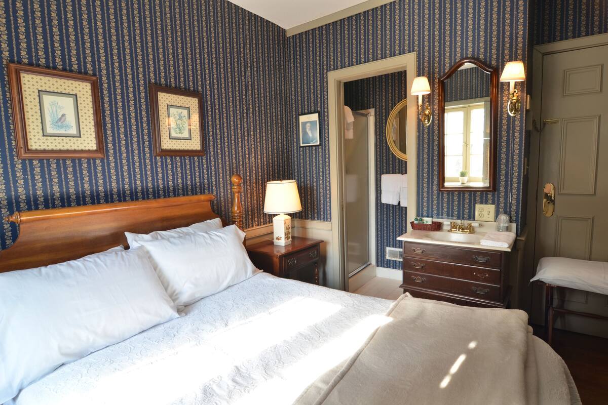 A hotel room with a large bed, a wooden headboard, a nightstand with a lamp, a dresser with a mirror, and a bathroom visible through an open door.