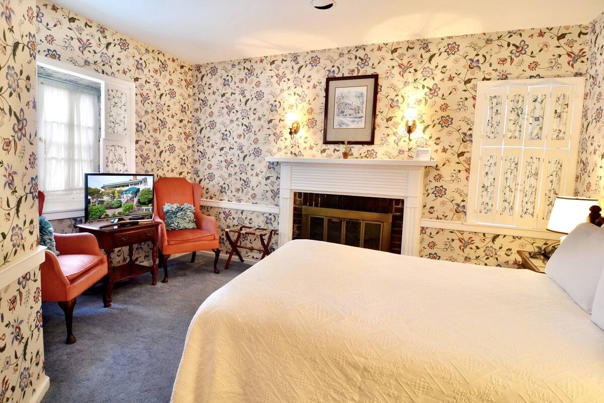 A hotel room with floral wallpaper, a fireplace, a bed, a desk with a television, and a window with curtains.