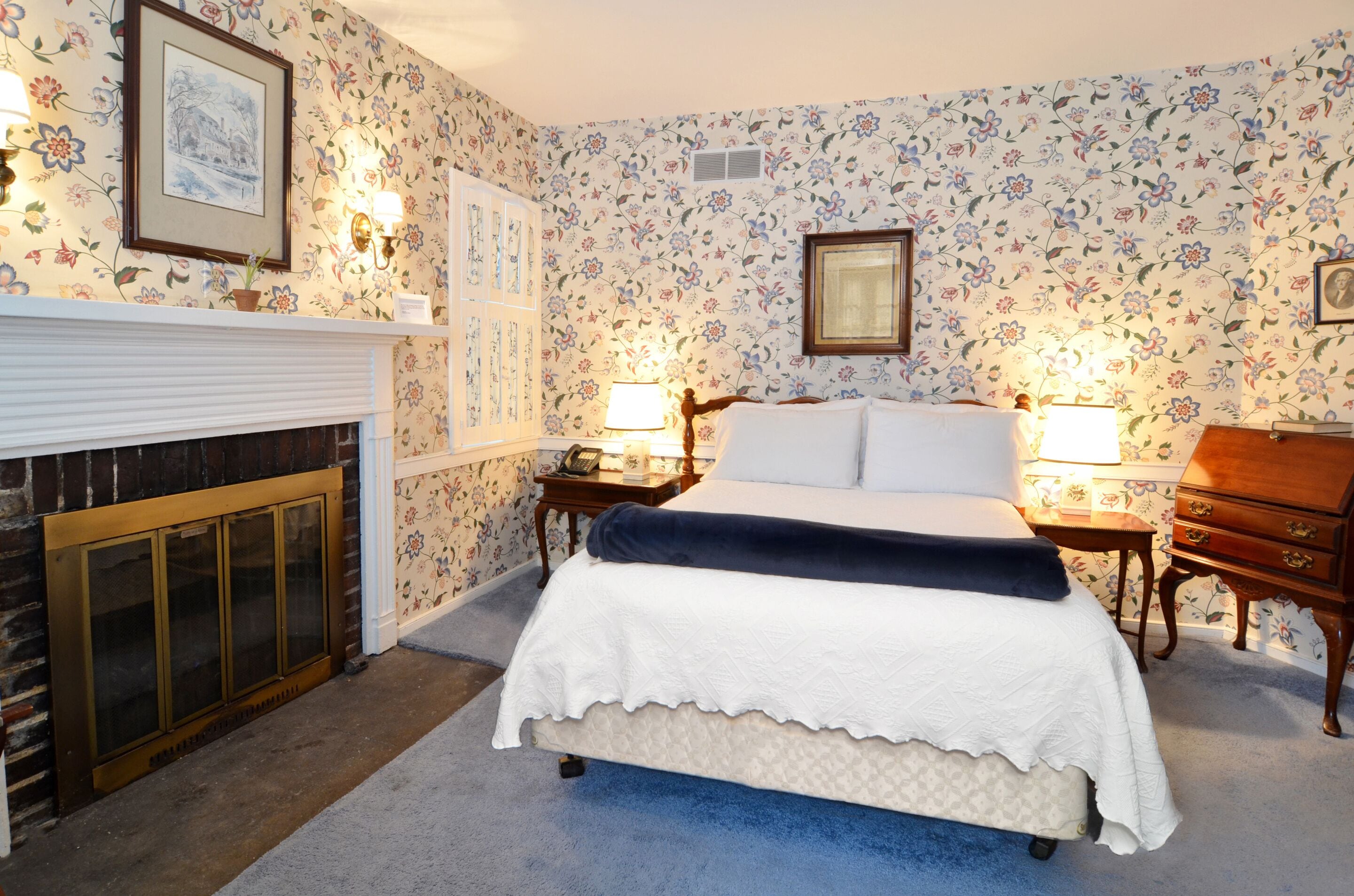 A bedroom with a bed, two bedside tables with lamps, a dresser, a fireplace, and floral wallpaper.