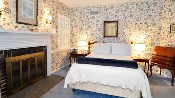 A bedroom with a bed, two bedside tables with lamps, a dresser, a fireplace, and floral wallpaper.