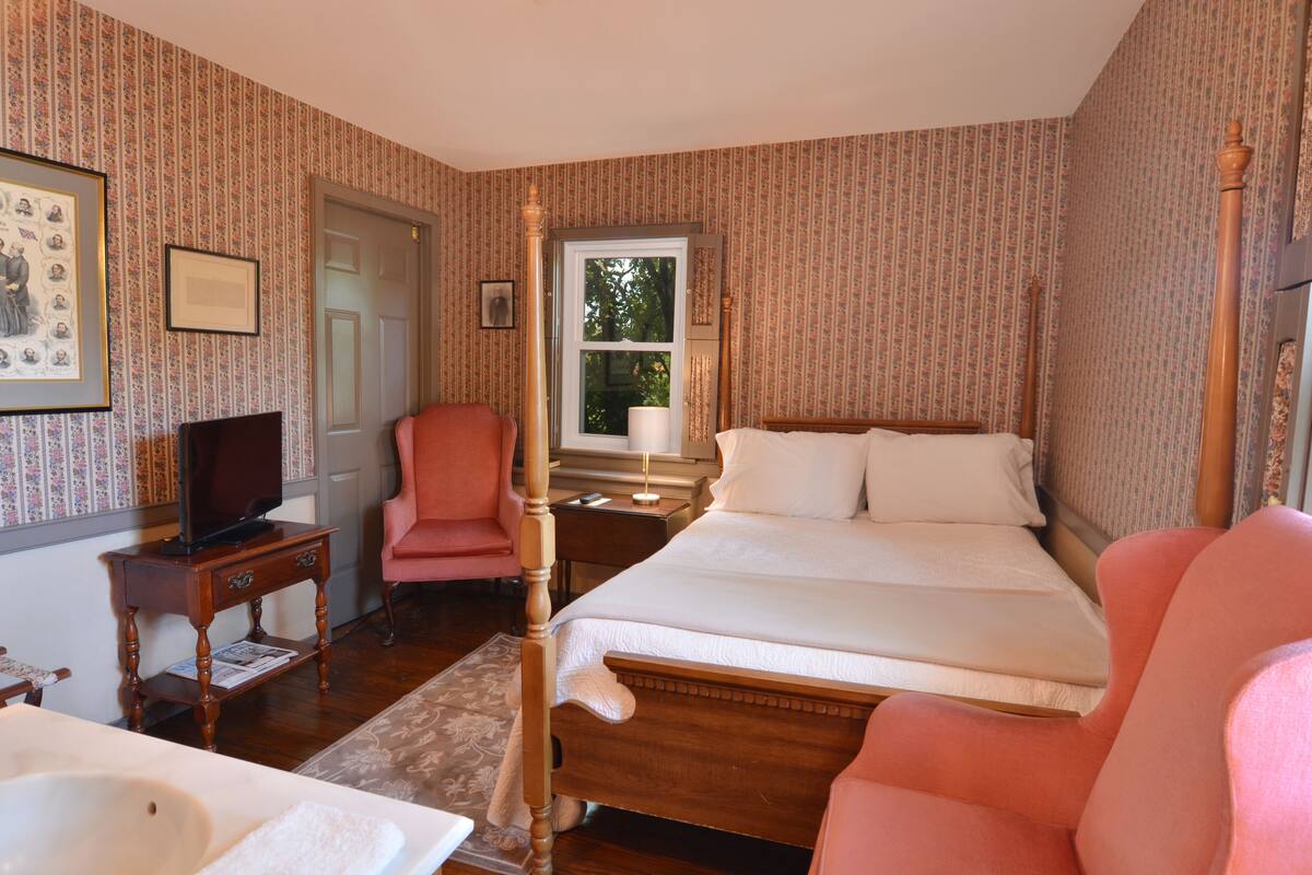 A bedroom with a wooden bed, a television on a side table, a chair, and a window with a view of trees.