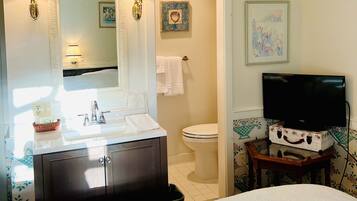 Un baño de hotel con lavamanos, espejo, inodoro y un televisor sobre una mesita.