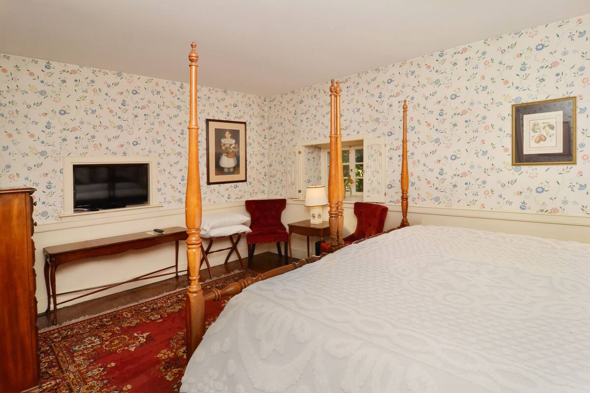 A four-poster bed with a red carpet, a wooden desk, a red chair, and a television on the wall.