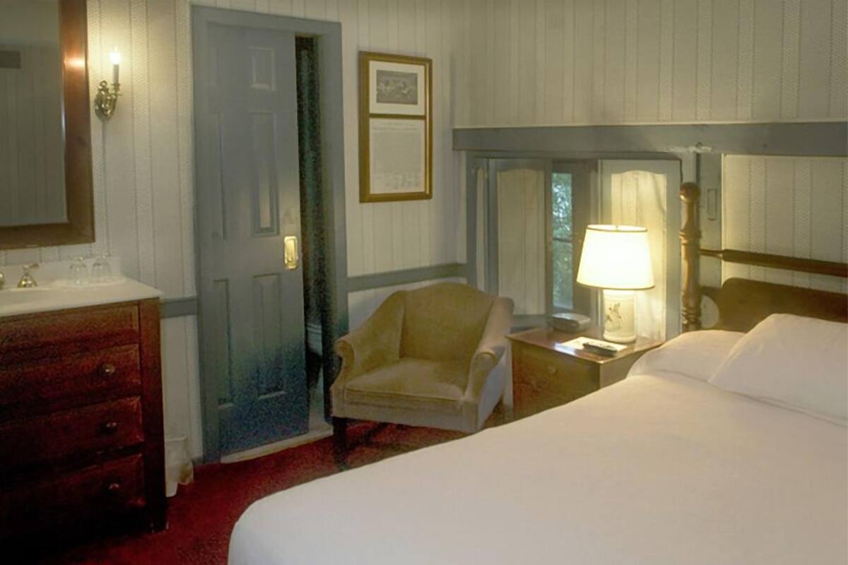 A hotel room with a bed, a chair, a nightstand with a lamp, a dresser, and a door.