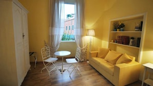 A room with a yellow wall, a white sofa, a small round table, two white chairs, a bookshelf, and a window with curtains. - Domus Ripetta (Rome)