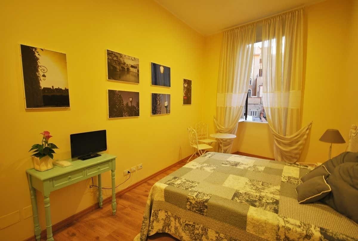 A room with a bed, a desk with a TV, a small table, and a chair. There are four framed pictures on the wall.