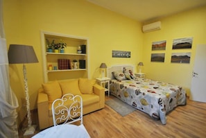 Una habitación con pared amarilla, una cama con colcha estampada, un sofá amarillo, una estructura de cama metálica blanca, una mesita de noche blanca con lámpara, un armario blanco, y una estantería con libros y objetos decorativos.