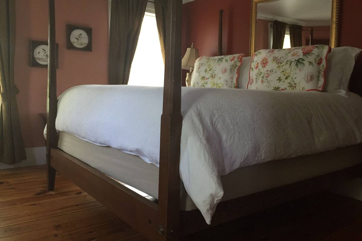 A four-poster bed with a floral bedspread, a wooden floor, a mirror with a gold frame, and a wall clock.