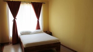 A single-bed room with a wooden frame, a bedside table, and a window with red curtains tied back. - Hospedaje Altamira (Florencia)