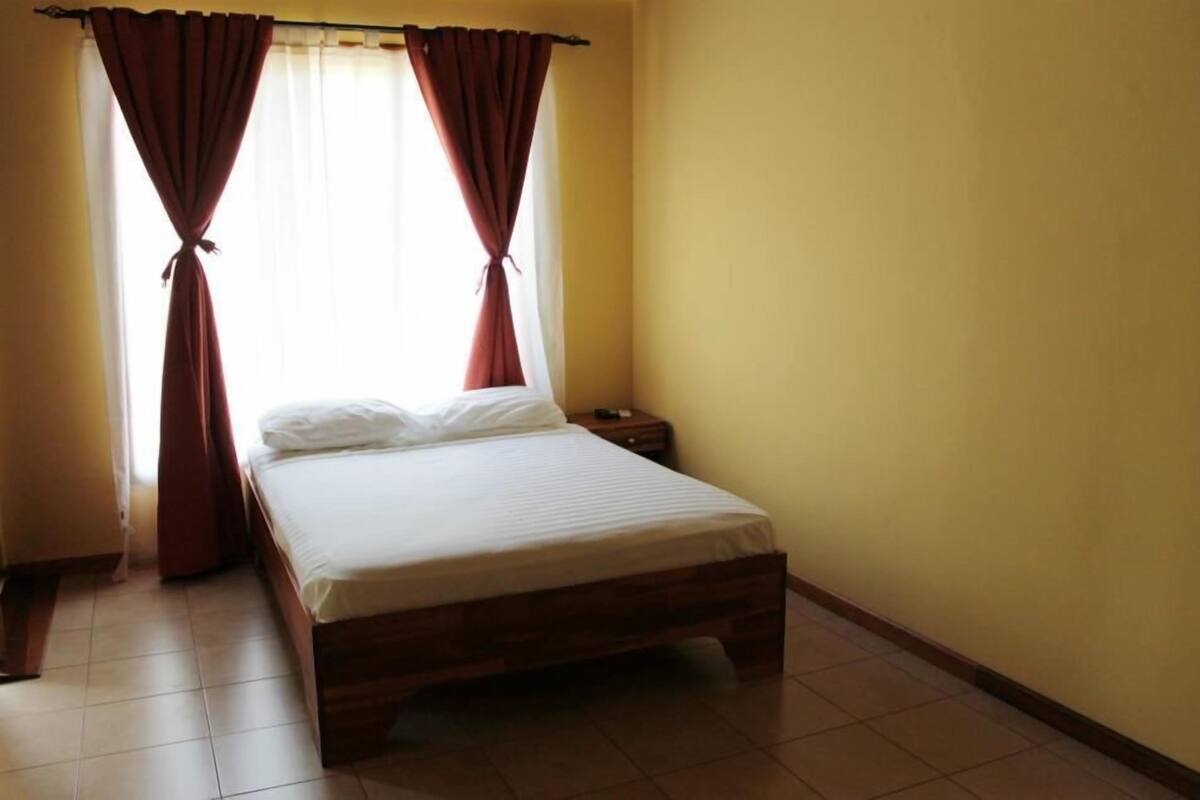 A single-bed room with a wooden frame, a bedside table, and a window with red curtains tied back.