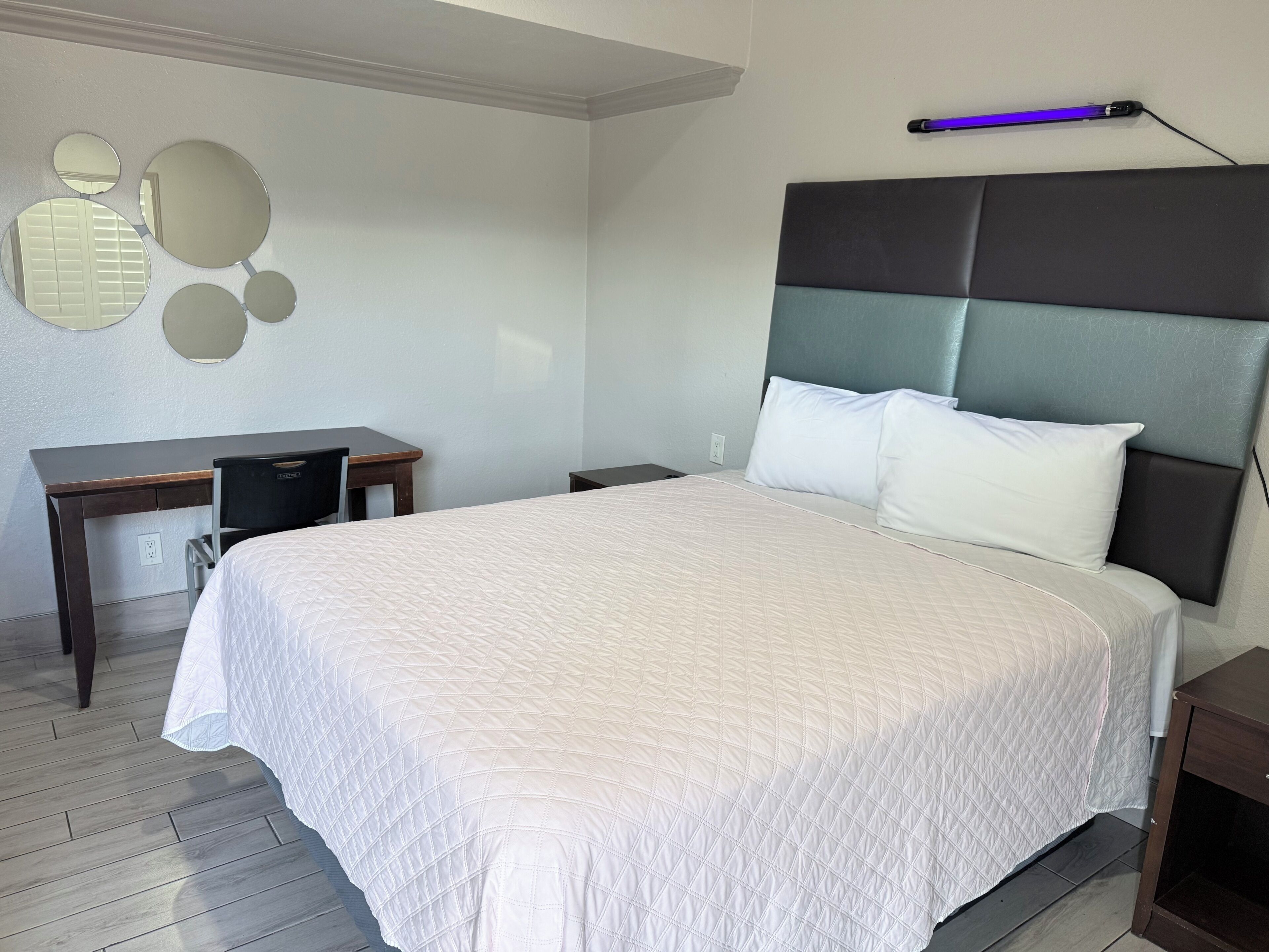 A hotel room with a large bed, a desk, a chair, a nightstand, and a wall-mounted lamp.