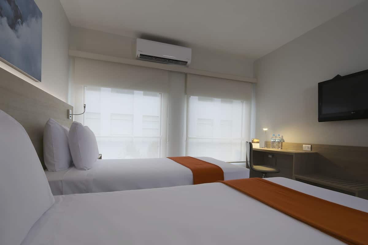 A hotel room with two beds, a desk, a chair, a TV, and an air conditioning unit.