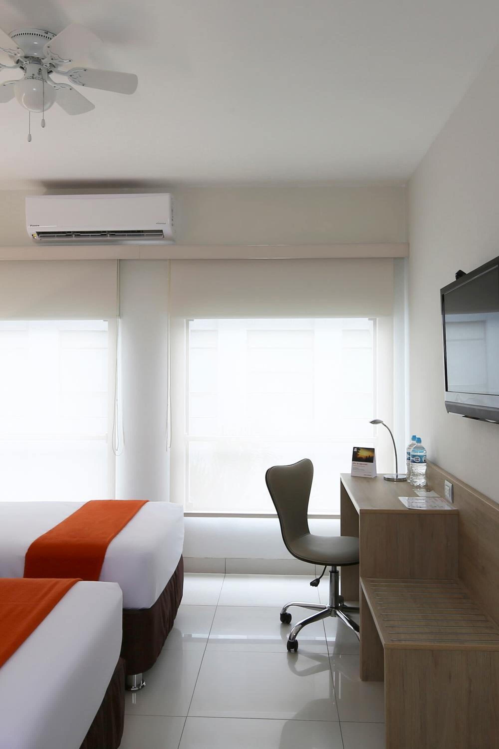A hotel room with two beds, a desk, a chair, a TV, and a ceiling fan.