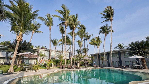Una piscina rodeada de palmeras y sillas de descanso, con un edificio al fondo.