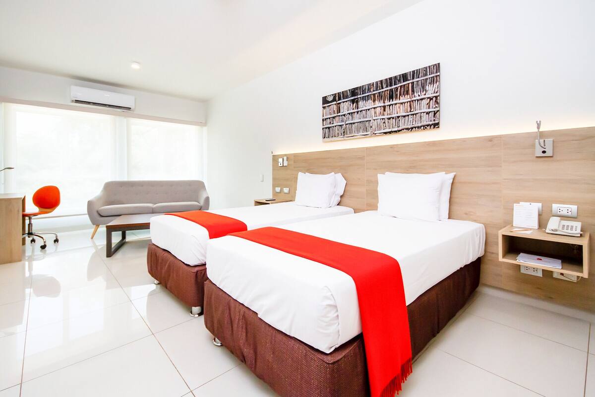 A hotel room with two beds, a desk, a chair, a bookshelf, and a large window.