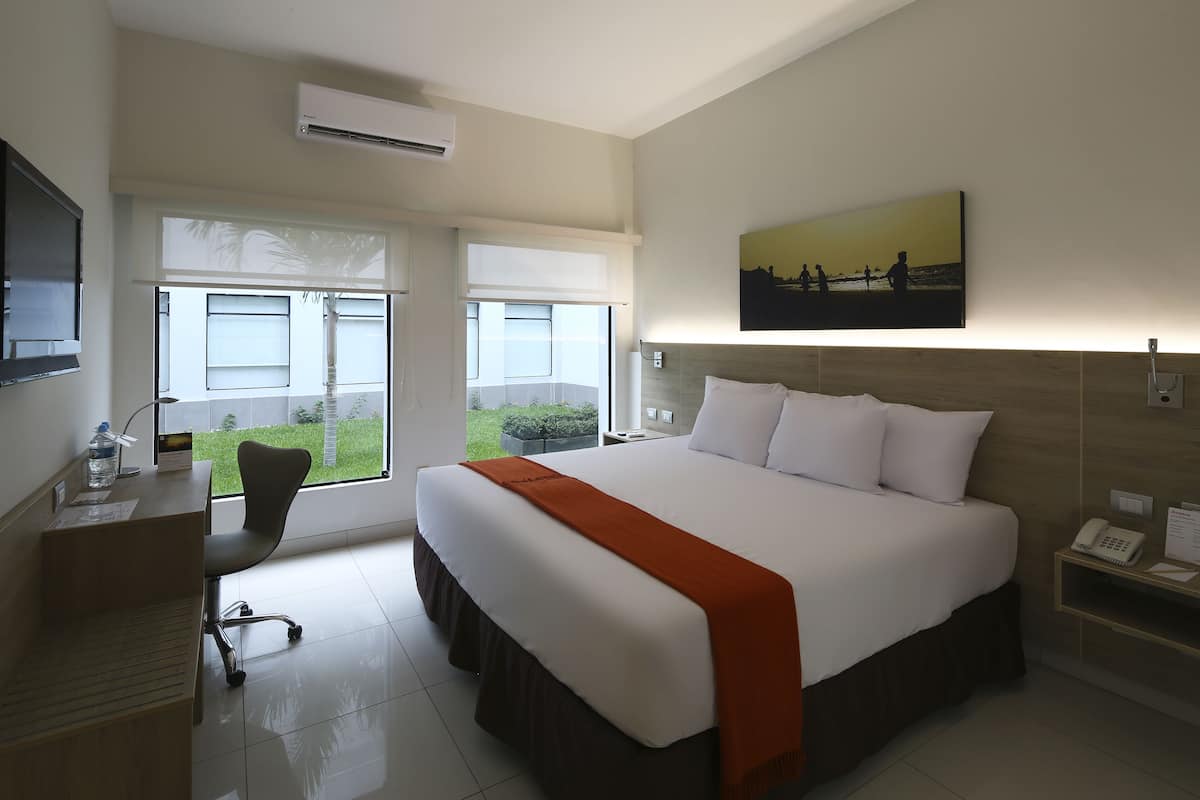 A hotel room with a large bed, a desk, and a chair. There is a window with a view of greenery outside.