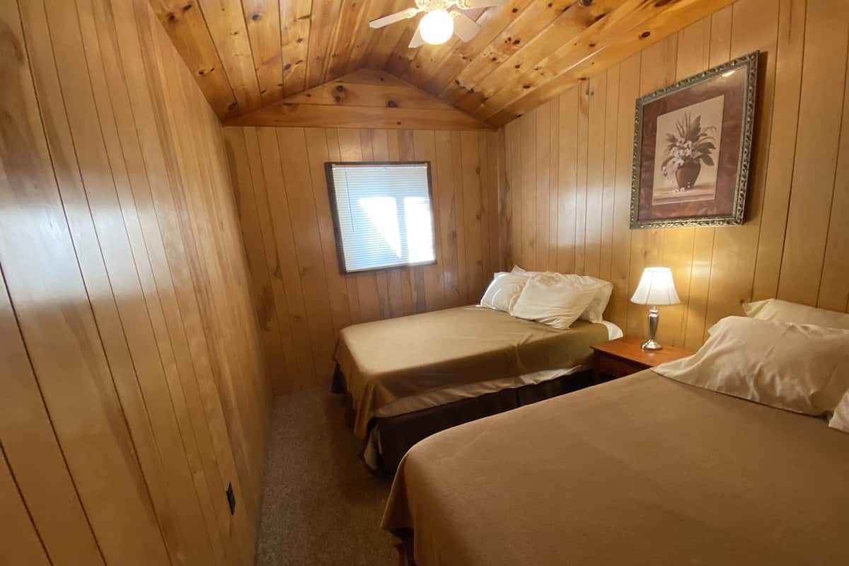 Une chambre rustique en bois avec deux lits, une fenêtre, un ventilateur de plafond et un tableau encadré au mur.