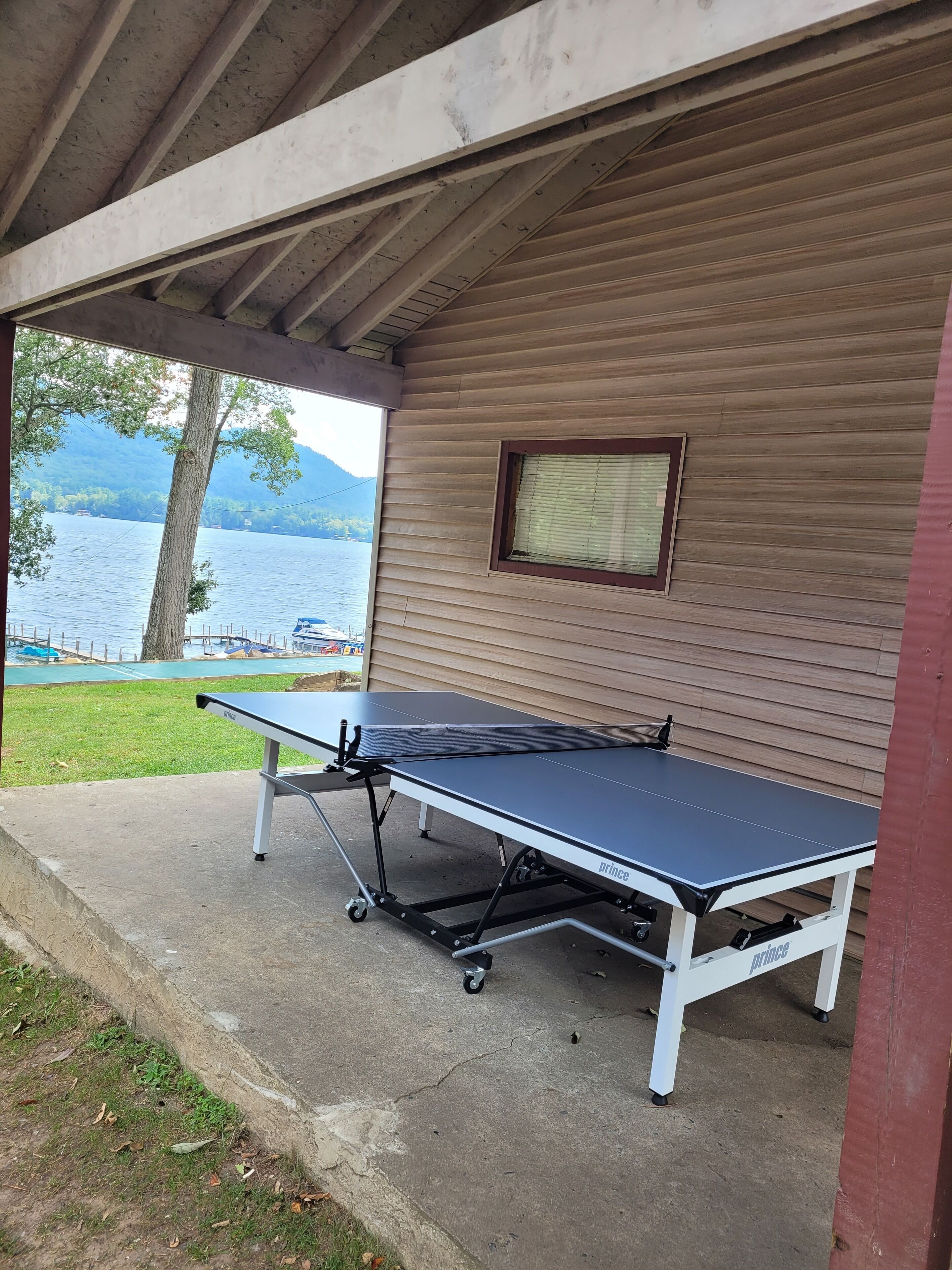 A ping pong table set up on a concrete surface under a covered area with a wooden wall and a window.