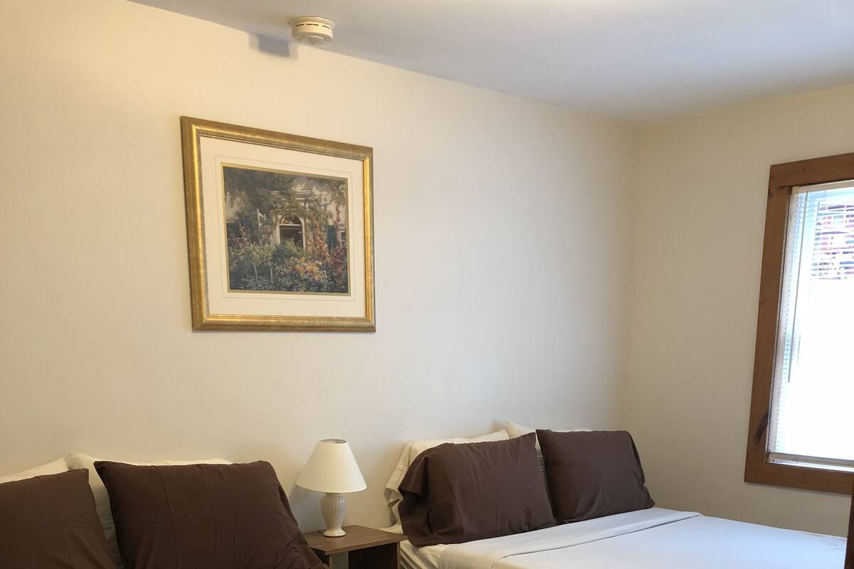 Une chambre d’hôtel avec deux lits, une table de chevet avec une lampe, un tableau encadré au mur et une fenêtre avec des stores.