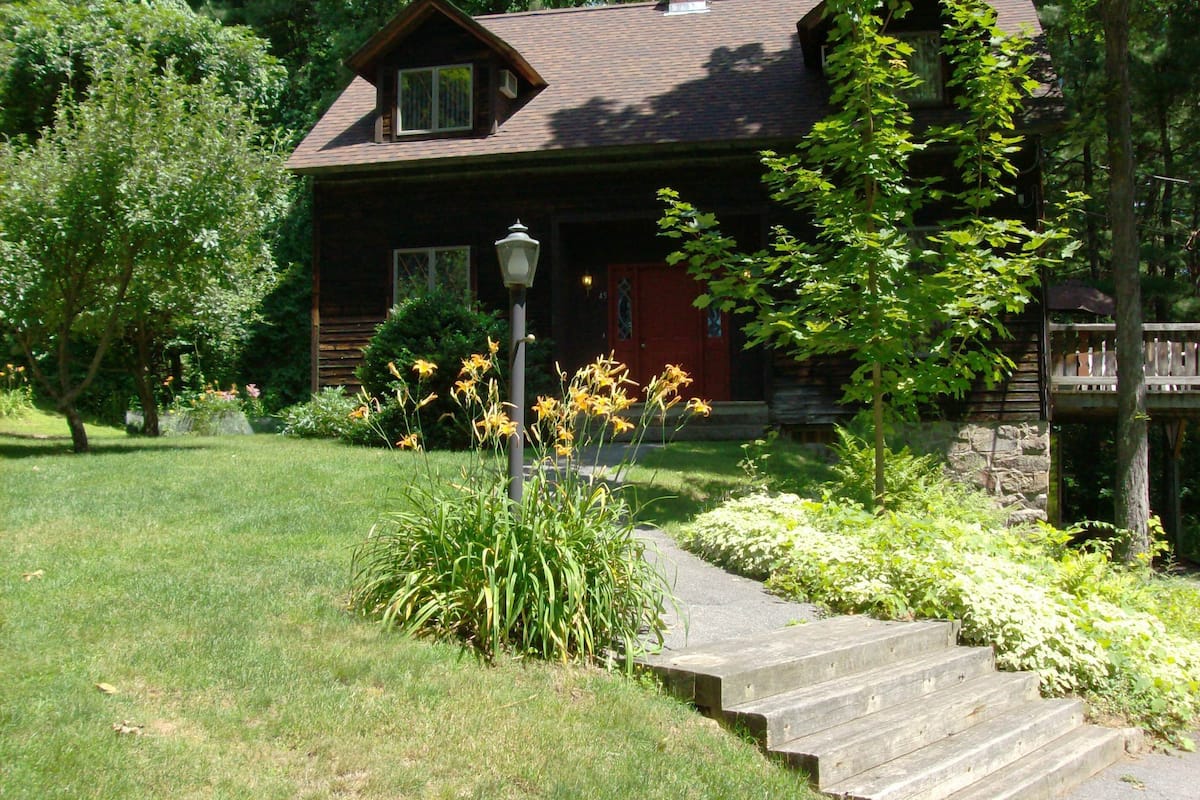 Une cabane en bois avec une porte rouge, entourée d’une végétation luxuriante et un chemin de pierres menant à l ’entrée.