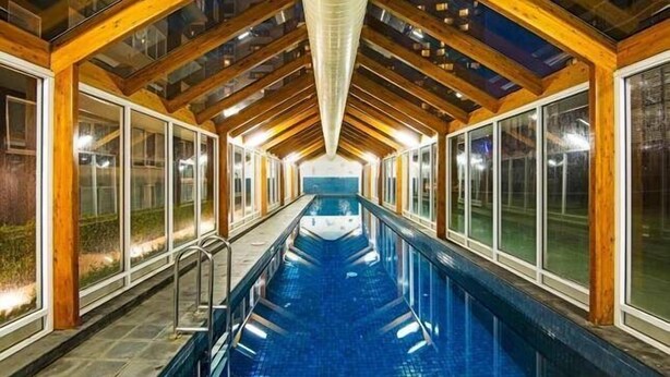 Una piscina interior con techo de cristal y vigas de madera, rodeada de grandes ventanales.
