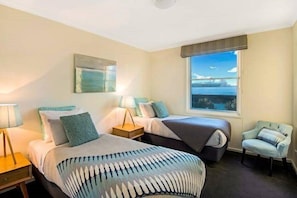 A hotel room with two beds, a window with a view, a chair, and a lamp. - Platinum Apartments on Whiteman St (Southbank)