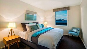Un dormitorio moderno con una cama grande, dos lámparas de noche, vistas de la ciudad a través de una ventana y una silla pequeña.