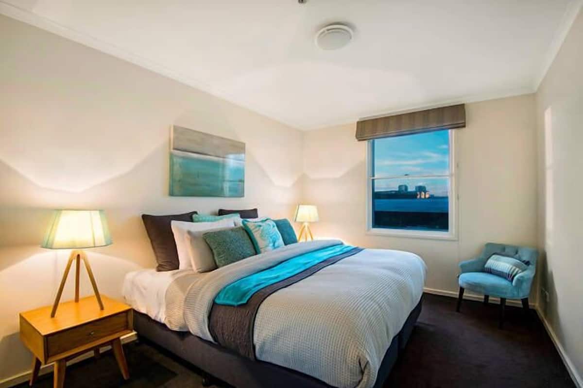 A modern bedroom with a large bed, two bedside lamps, a view of the city through a window, and a small chair.