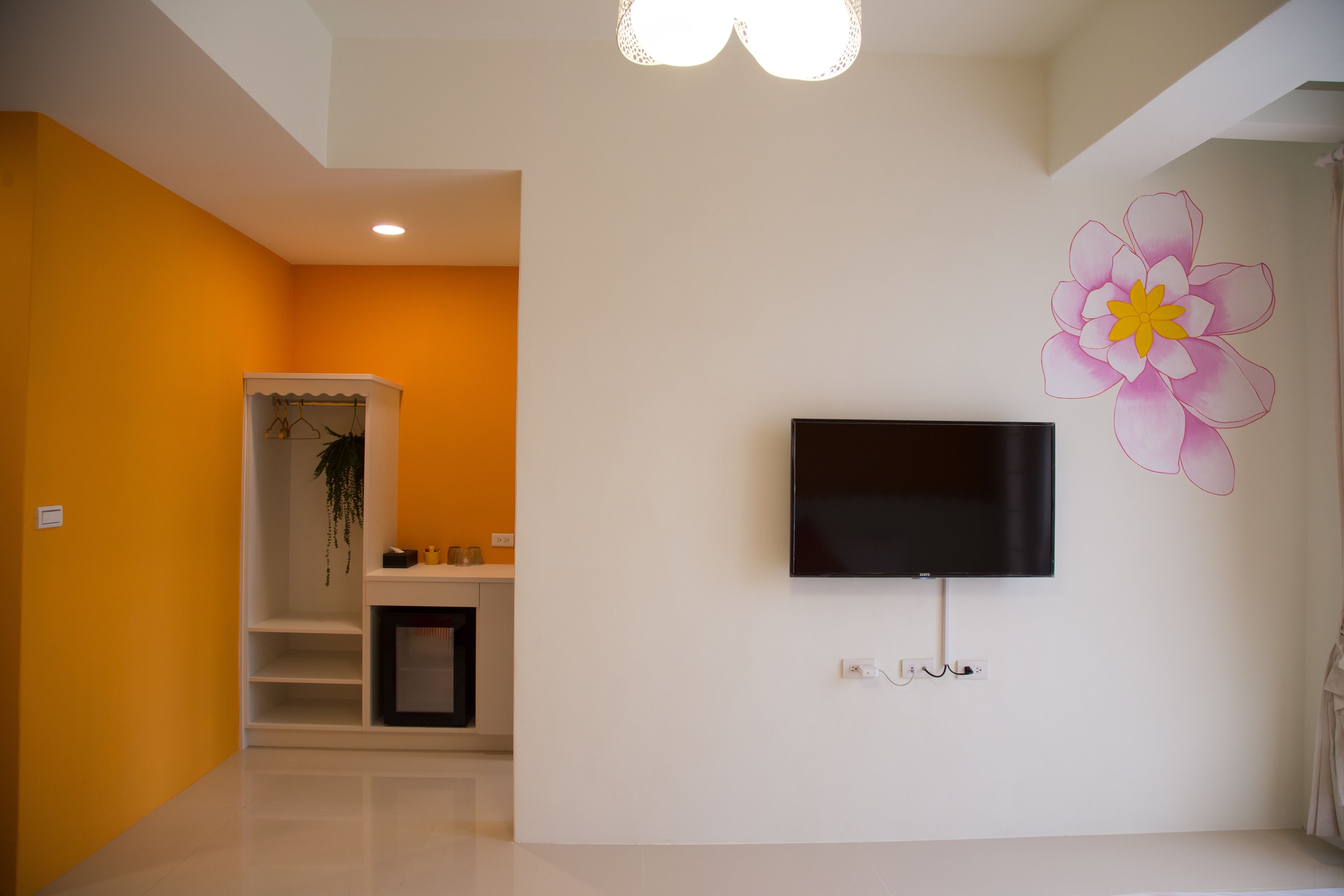 A modern living room with a flat-screen TV mounted on the wall, a large flower mural, and a white shelving unit with a hanging plant.