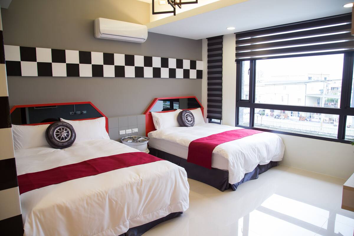 A hotel room with two beds, checkered wall decor, a large window with a city view, and an air conditioning unit.