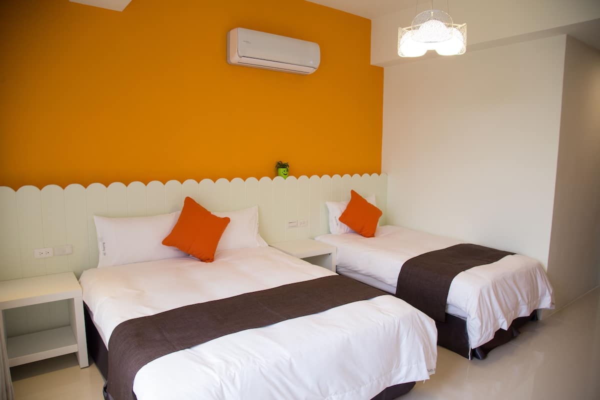 A hotel room with two beds, white bedding, orange pillows, a white nightstand, an air conditioning unit, and a white ceiling light.