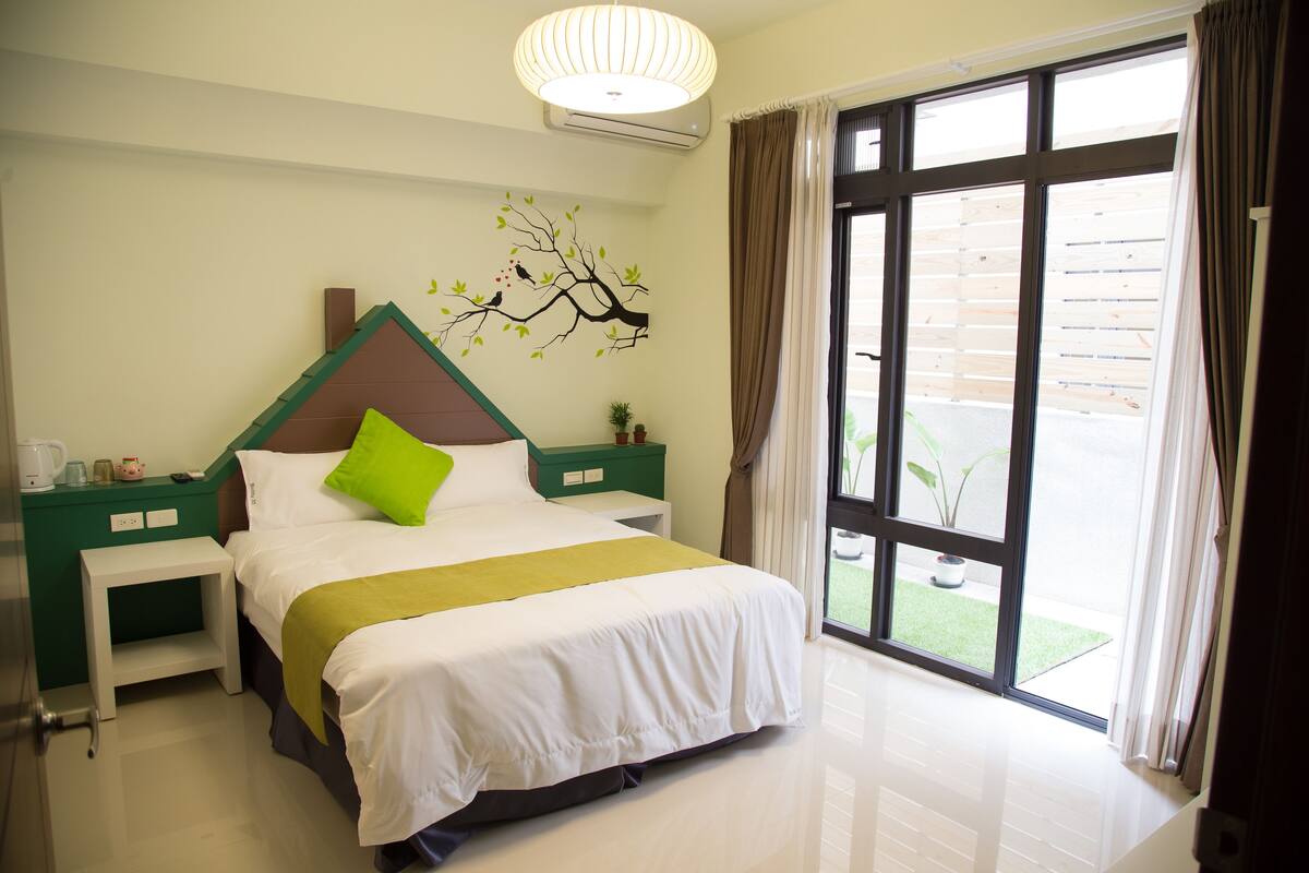 A hotel room with a bed, green nightstands, a white lamp, and a window with curtains.
