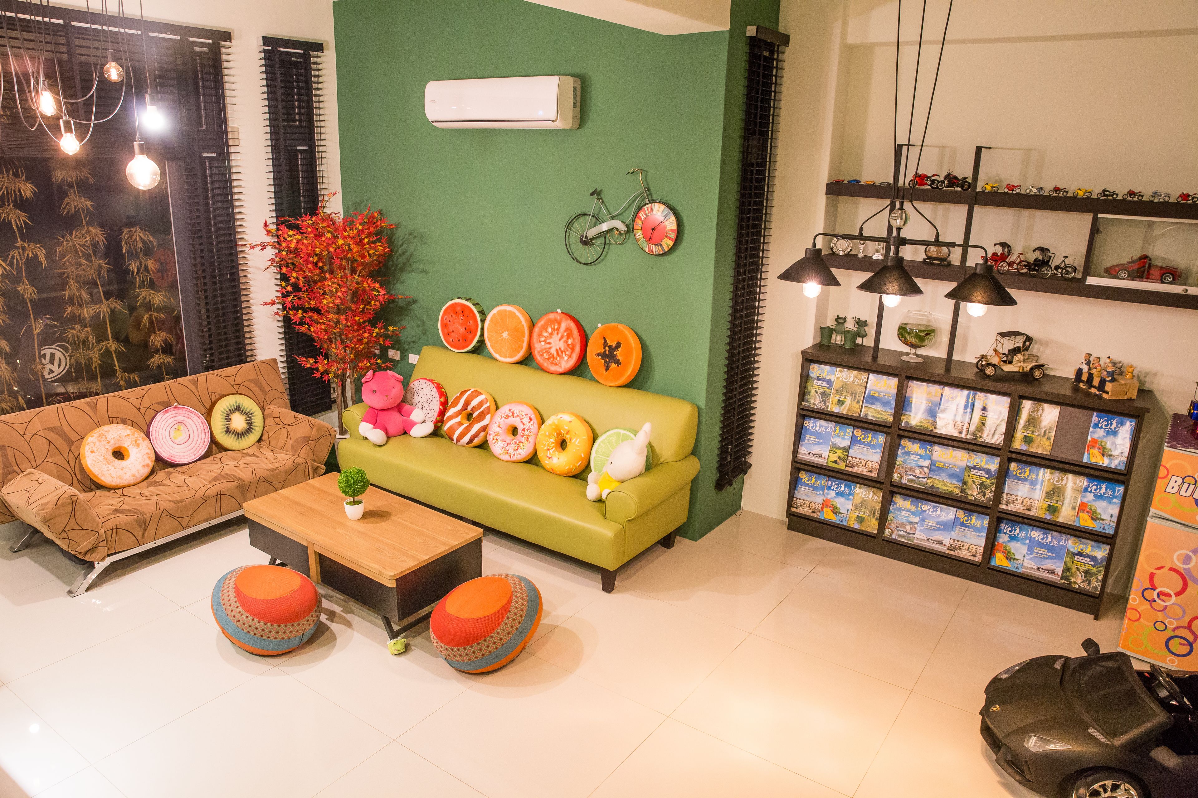 Una sala de estar moderna con una pared verde, un sofá marrón con cojines de forma toroidal, un sofá verde con cojines con forma de fruta, una mesita y una estantería llena de libros de viajes.