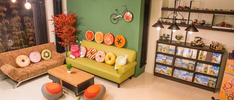 Una sala de estar moderna con una pared verde, un sofá marrón con cojines de forma toroidal, un sofá verde con cojines con forma de fruta, una mesita y una estantería llena de libros de viajes.