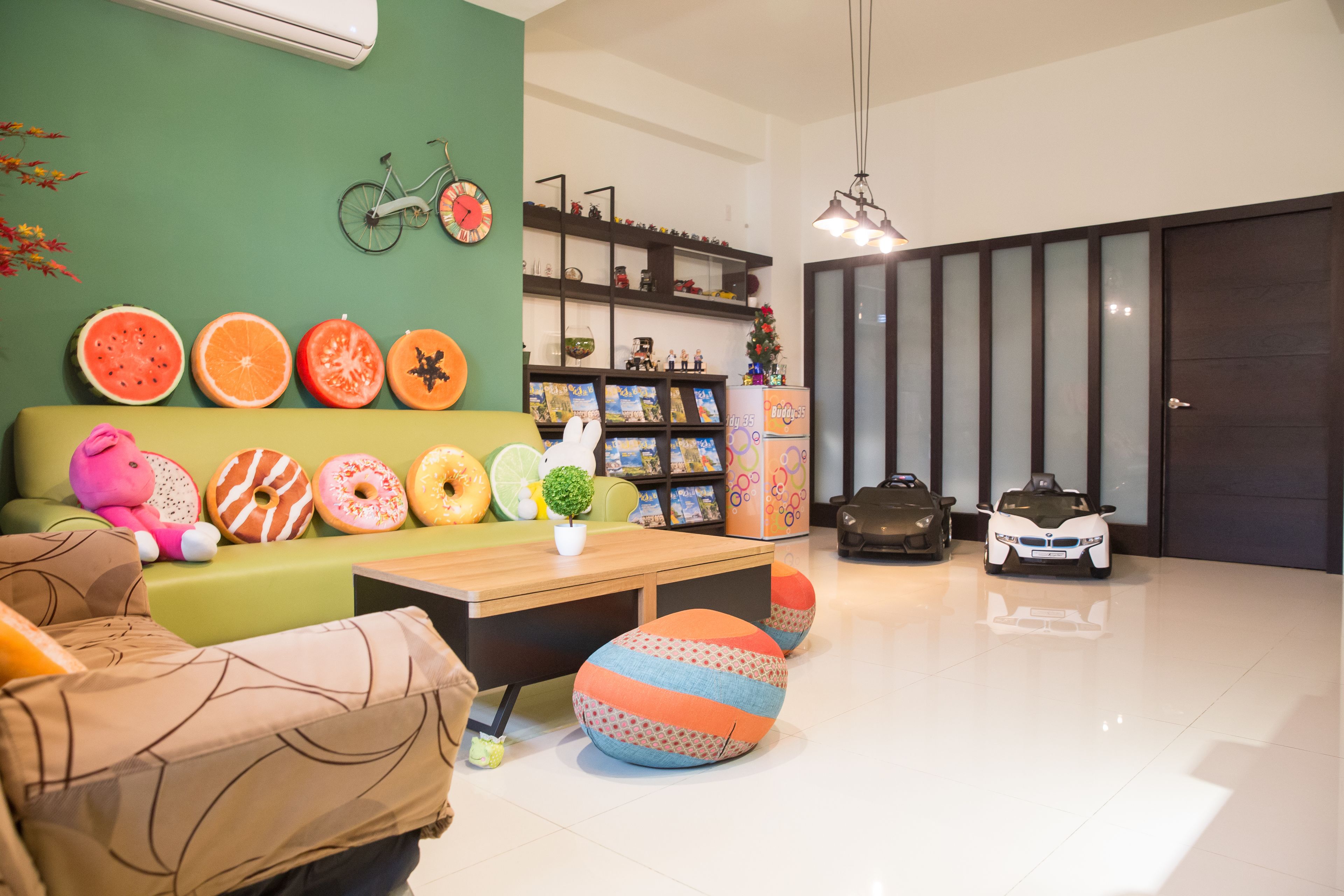Una sala de estar moderna con un sofá verde, una mesa de centro de madera y una variedad de objetos coloridos, incluyendo una bicicleta, un reloj, y una colección de libros y juguetes.