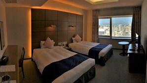Een hotelkamer met twee bedden, een bureau met stoel, een kleine tafel, een televisie en een raam met uitzicht op de stad.