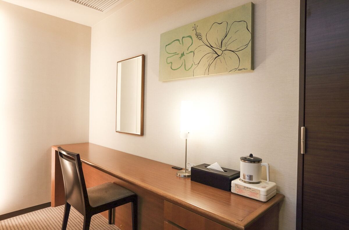 A hotel room with a wooden desk, a chair, a bedside table with a lamp, a tissue box, and a coffee maker.