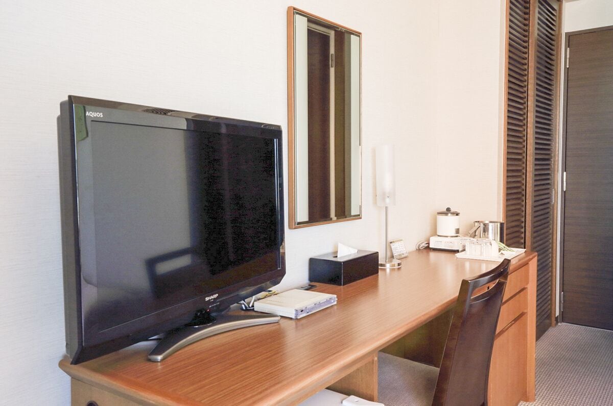 A hotel room with a wooden desk, a flat-screen TV, a mirror, a bedside table with a lamp, and a door with a sliding shutter.
