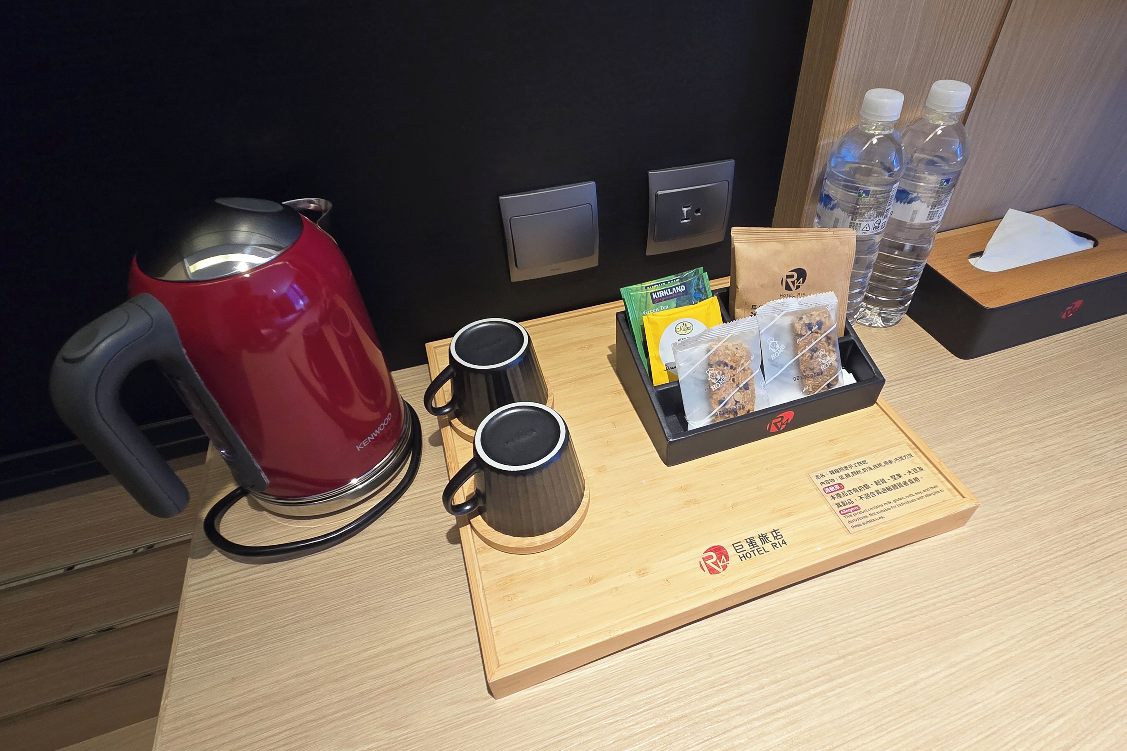 Ein Hotelzimmer mit einem Holftablett, das einen roten Wasserkocher, zwei schwarze Tassen, eine Taschentuchbox, eine kleine Tasche und zwei Wasserflaschen enthält.