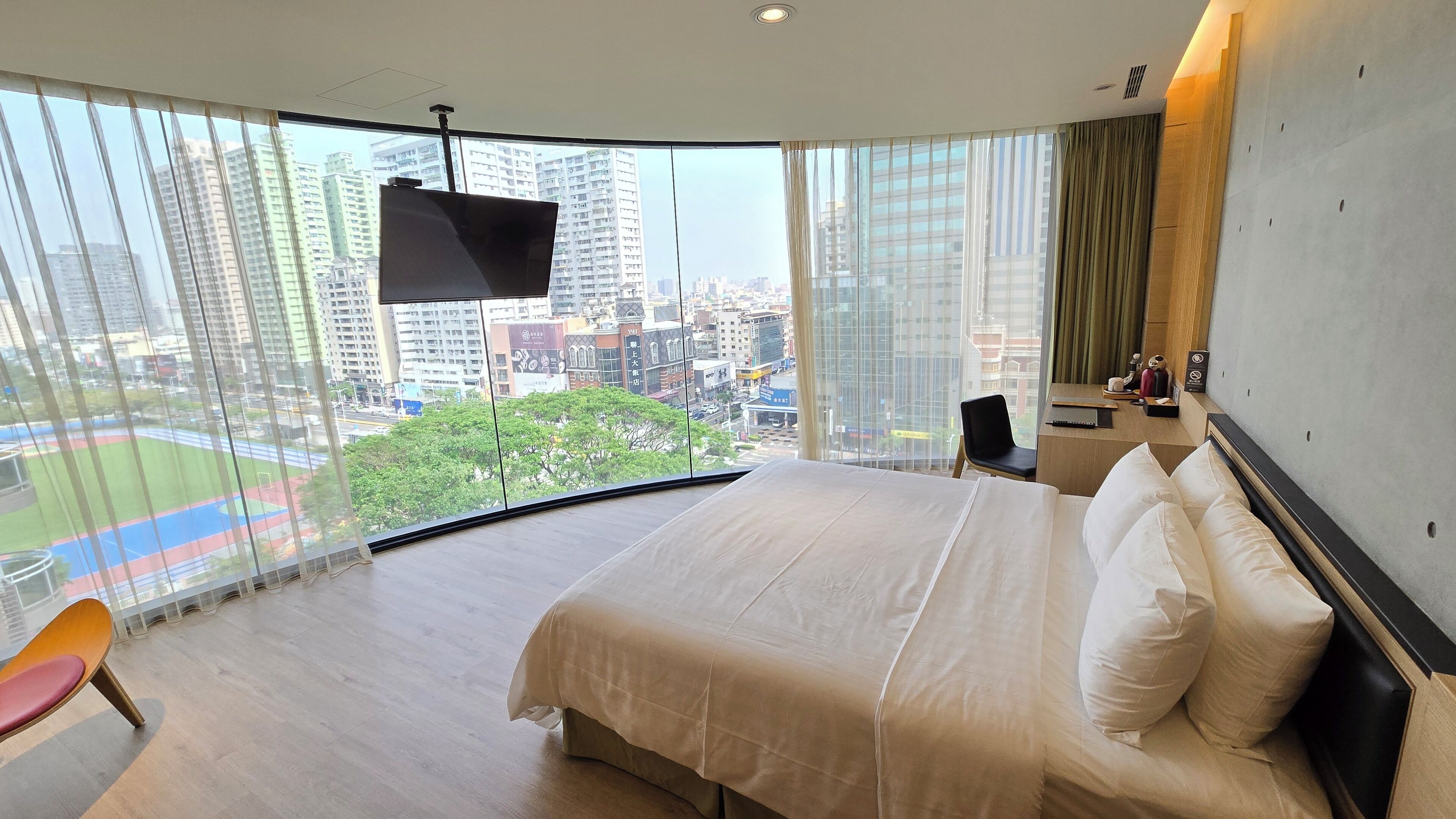 A modern hotel room with a large bed, a desk, and a view of a cityscape through floor-to-ceiling windows.