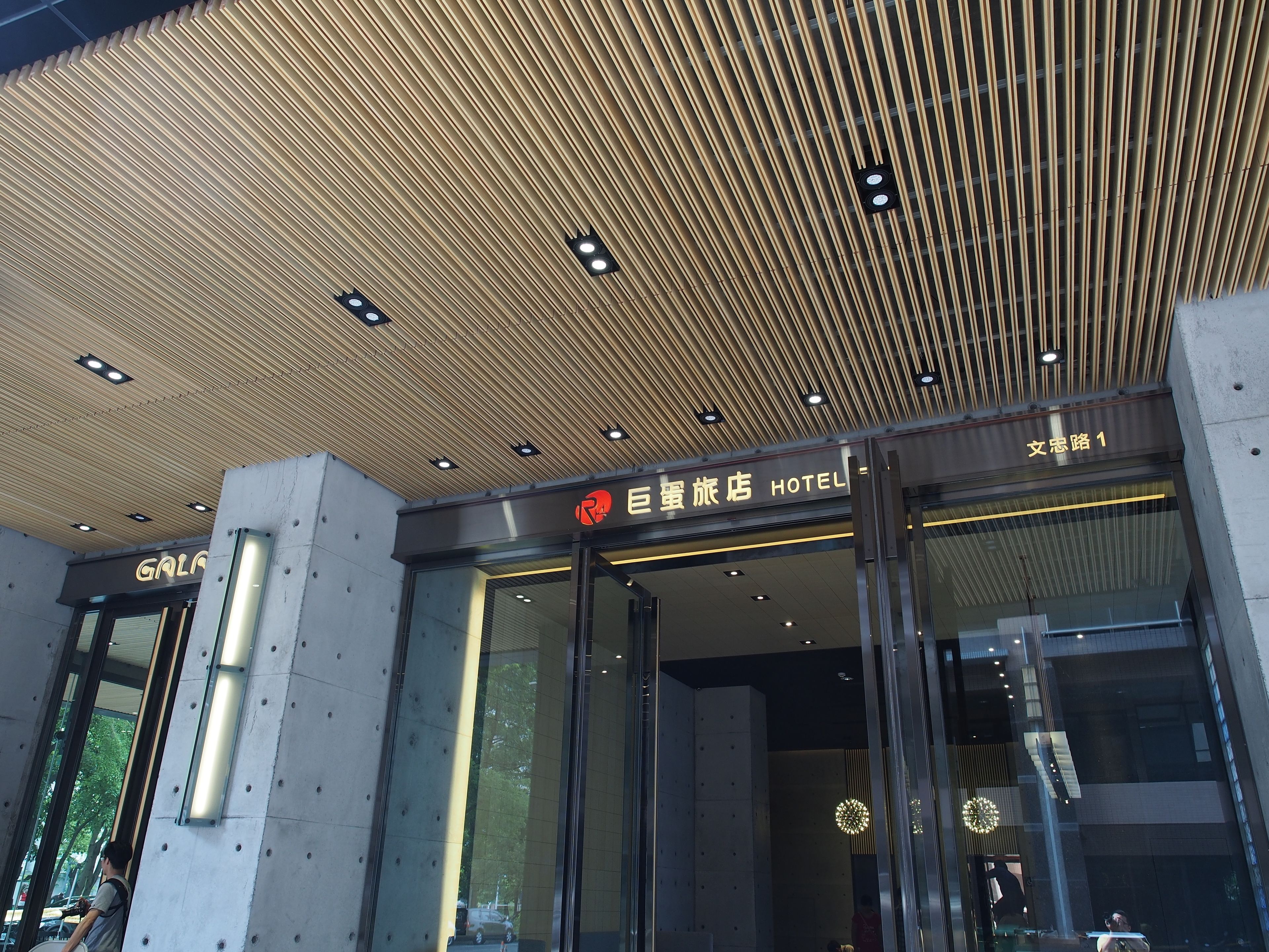 Entrada de hotel con diseño moderno, destacando un techo de listones de madera y puertas de vidrio.